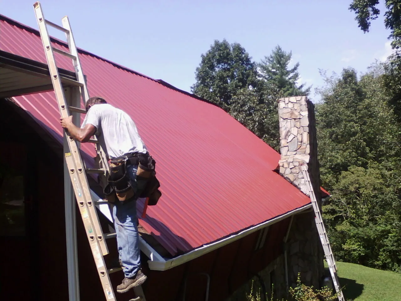 Roofer on a ladder beside a red metal roof and stone chimney — Soffit & Fascia Repair by Roofing Brothers in Shadyside